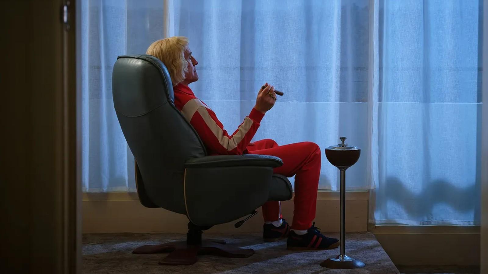Steve Coogan sitting in a chair smoking a cigar as Jimmy Savile in The Reckoning.